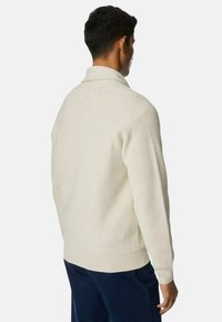 Helle beige Strickpullover mit geripptem Muster, ausgestattet mit einem umgeschlagenen Kragen und langen Ärmeln, getragen mit dunkelblauen Hosen.