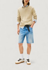 Beżowy bawełniany sweter noszony na jasnoniebieskiej koszuli w paski, zestawiony z dżinsowymi szortami i białymi trampkami. Obecny czarny pasek oraz detal postrzępionych szortów.