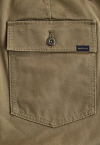 Taschini in tessuto beige con chiusura a bottone e bordi cuciti, dotato di una piccola etichetta blu sul lato che riporta la scritta "SUPPLY Co.".