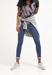 Svart t-shirt med grå Nike-logga, grå sweatshirt knuten runt midjan, blå skinny jeans och grå träningsskor med texturerade detaljer.