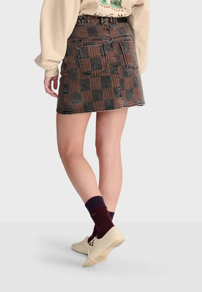 Femme portant une mini-jupe à carreaux marron et noire, des chaussures beiges et des chaussettes foncées, debout devant un fond clair uni.