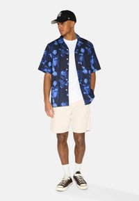 Short-sleeve, navy floral shirt with blue patterns, paired with light beige shorts and brown sneakers, topped with a black cap.