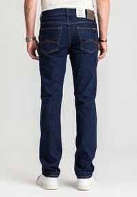 Ciemnoniebieskie jeansy z prosto wyciętą nogawką. Charakteryzują się fakturą powierzchni, żółtym przeszyciem oraz charakterystycznym wzorem tylnej kieszeni. Widoczne białe trampki.