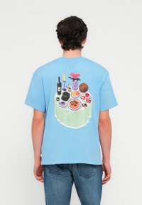 Ανοιχτό μπλε βαμβακερό t-shirt με έντονο γραφικό στο πίσω μέρος, που απεικονίζει ένα τραπέζι με φαγητό, λουλούδια και διακοσμητικά αντικείμενα σε λεπτομερείς αποχρώσεις.