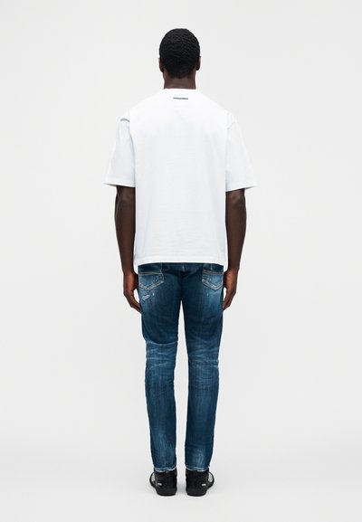 Λευκό βαμβακερό μπλουζάκι με κοντά μανίκια, εμφανιζόμενο από πίσω. Συνδυάζεται με φθαρμένο μπλε τζιν και μαύρα αθλητικά παπούτσια.