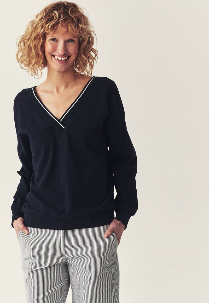 Femme souriante aux cheveux blonds bouclés portant un haut noir à manches longues avec col en V et un pantalon gris clair rayé, debout les mains dans les poches.