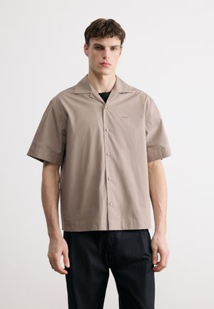 Ung mann iført en beige kortermet skjorte knapper foran over en svart topp, kombinert med svarte bukser, stående mot en ensfarget hvit bakgrunn.