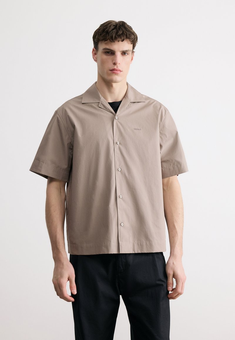 Jeune homme portant une chemise beige à manches courtes boutonnée par-dessus un haut noir, associé à un pantalon noir, debout devant un fond blanc uni.