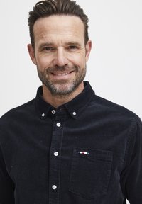 Svart kordfløyels skjorte med knapp, med en lomme som har et lite rødt, hvitt og blått akcent. Hvite knapper og en klassisk kragedesign.