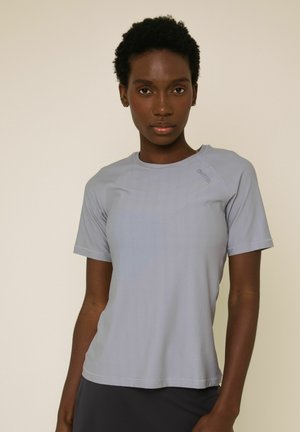 Femeie purtând un tricou sportiv ușor gri deschis cu mâneci scurte și pantaloni închiși la culoare, stând în picioare pe un fundal bej simplu.