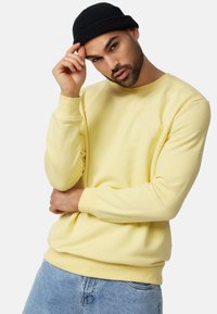 Lys gul bomulds sweatshirt med rund hals, ribbede manchetter og kant, med et lille logo på brystet, bæres sammen med blå denimjeans.