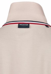 Polo beige avec un col haut, une touche à rayures noires et blanches, et une étiquette portant la mention "HECHTER PARIS" située à l'arrière.