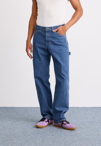 Blå denim jeans med rett snitt, med en klassisk fem-lomme-design og lyse slitasjedetaljer. Bæres med lilla sko.