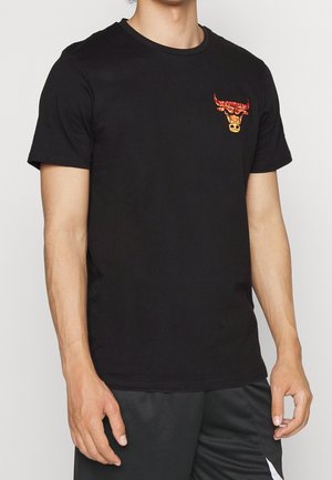 Homme portant un t-shirt noir avec un petit motif de tête de taureau rouge et orange sur le côté gauche de la poitrine, assorti à un short noir.