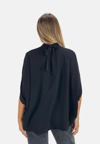 Schwarze Bluse mit hochgeschlossenem Kragen und Schleifen-Detail am Rücken. Lässige Passform mit kurzen Ärmeln, aus leichtem, strukturiertem Stoff gefertigt.