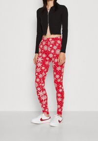 Rode leggings met witte sneeuwvlokpatronen, gecombineerd met een zwart kort jack met rits en witte sneakers met een rood swoosh-logo.