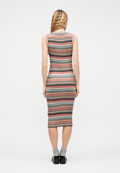 Femme debout, de dos, portant une robe tricotée rayée sans manches, longueur genou, des chaussettes grises et des chaussures noires.