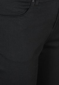 Mustade teksade lähivõte, kus on nähtav eesmine tasku, õmblused ja nööp sulgemisel.