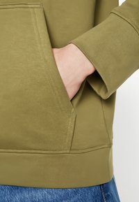 Mano inserita nella tasca anteriore di una felpa verde oliva, che mostra i dettagli delle cuciture e dei polsini, con i pantaloni blu testurizzati visibili sotto.