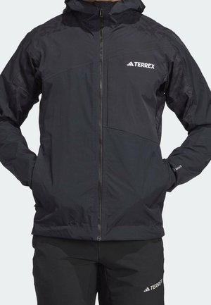 Persona con chaqueta con capucha negra de la marca Terrex y pantalones a juego con las manos en los bolsillos de la chaqueta sobre un fondo liso.
