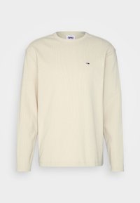 Beżowa koszulka z długim rękawem, wykonana z prążkowanej tkaniny, z okrągłym dekoltem i małym haftowanym logo w kolorach czerwonym, białym i niebieskim na piersi.