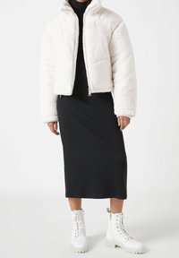 Chaqueta corta de piel sintética blanca con cierre de cremallera, combinada con un vestido midi negro de canalé y botas de combate blancas con cordones.
