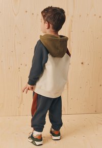 Mikina s farebnými blokmi v zelenej, bielej a námorníckej modrej; v kombinácii s námorníckymi teplákmi s oranžovými bočnými pruhmi; tenisky s gumovou podrážkou.