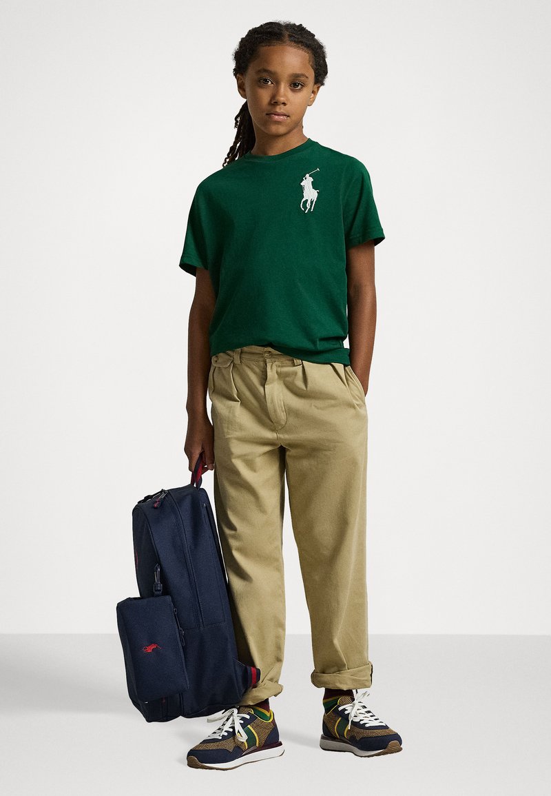 Grünes Baumwoll-T-Shirt mit weißem Logo, beigefarbene, plissierte Hose, marineblauer Rucksack mit roten Akzenten und mehrfarbige Sneakers mit weißen Sohlen.