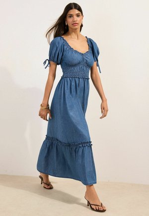 Frau im Mittelschritt trägt ein blaues Kleid mit Puffärmeln, geraffter Taille und Rüschensaum, kombiniert mit schwarzen Sandalen mit Absatz und goldenen Armbändern.