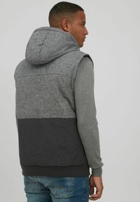 Graue Kapuzenweste mit einem mehrschichtigen Design, das ein oberes Fischgrätenmuster, einen kontrastierenden einfarbigen dunklen unteren Abschnitt und gerippte Bündchen aufweist.