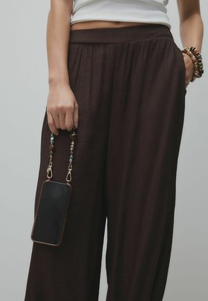 Brūnas, teksturētas plataji bikses ar elastīgu jostasvietu, nēsātas ar roku, kas tur viedtālruni piesaistītu pie pērlīšu siksniņas.