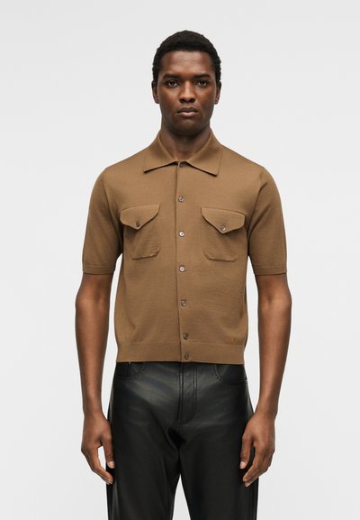 Άνδρας που στέκεται με το πρόσωπο προς τα εμπρός φοράει καφέ κοντομάνικο πουκάμισο με τσέπες στο στήθος και μαύρο δερμάτινο παντελόνι μπροστά σε απλό φόντο.