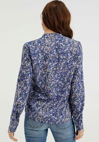 Chemise à manches longues avec un motif floral dans des teintes de violet, blanc et bleu. Présente un col froncé et une texture de tissu lisse.