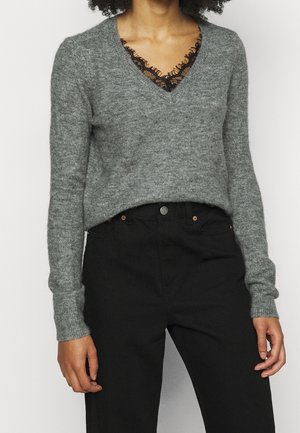 Persoon draagt een grijze V-hals gebreide trui over een zwarte top met kanten rand, gecombineerd met een zwarte broek met hoge taille.
