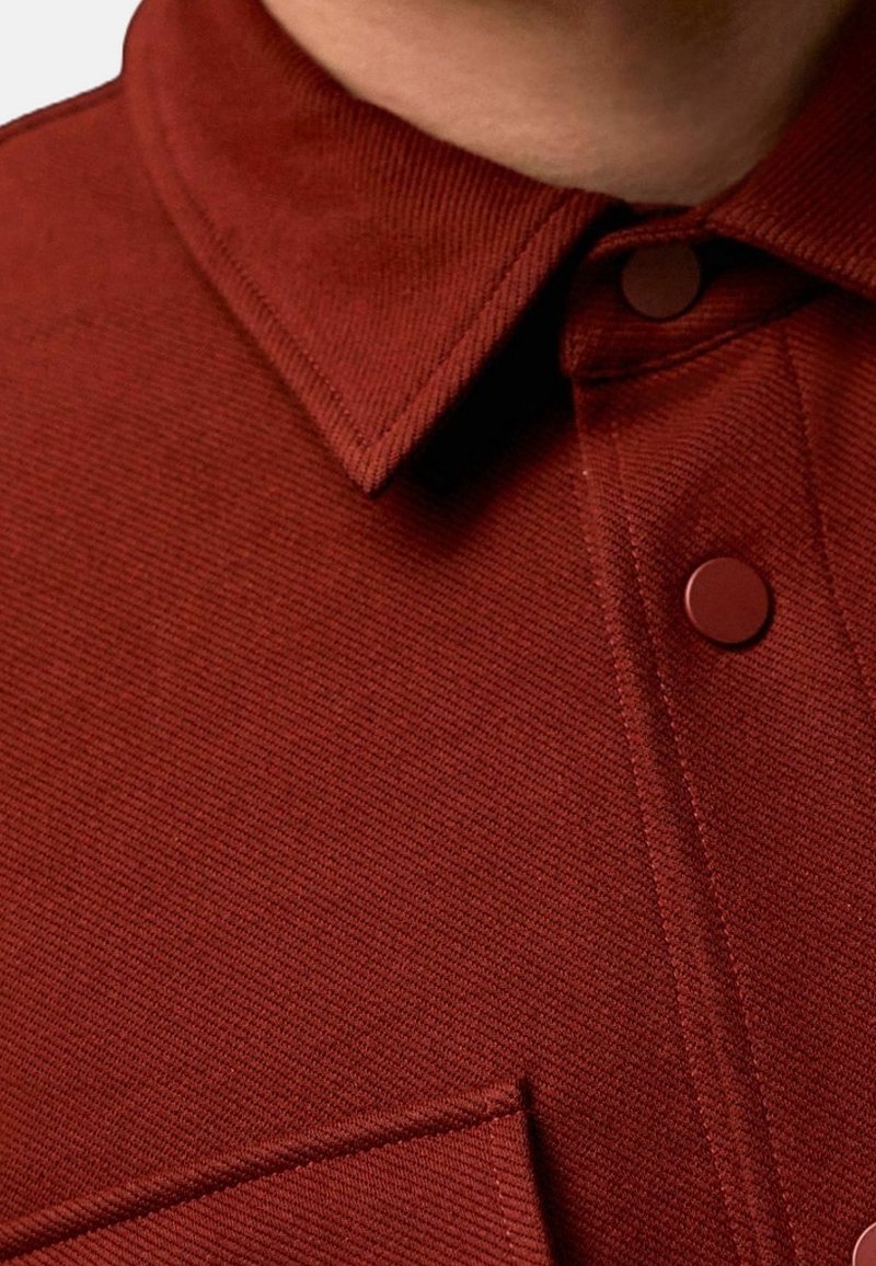 Gros plan sur le col d'une chemise couleur rouille avec des boutons-pression assortis et un tissu texturé.