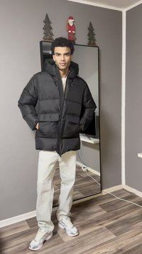 Zwarte pufferjas met capuchon, voorzien van een ritssluiting aan de voorkant en twee zakken aan de voorkant. Draagt over een beige top en witte broek met grijze sneakers.