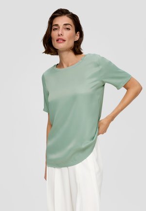 Blouse mint vert à manches courtes avec un col rond, texture lisse et ourlet légèrement incurvé, associée à un pantalon blanc.