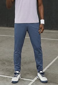 Sportowiec w niebieskich spodniach, białych butach sportowych i białej opasce na nadgarstek stojący na korcie tenisowym blisko linii granicznych.