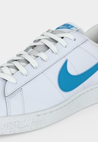 Λευκό αθλητικό παπούτσι Nike με μπλε λογότυπο swoosh, λευκά κορδόνια και υφή στη σόλα, εμφανίζεται από το πλάι και ελαφρώς από πάνω.