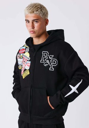 Jeune personne aux cheveux blonds courts portant un sweat à capuche noir zippé avec des patchs colorés et un motif de croix blanche sur la manche, les mains dans les poches.