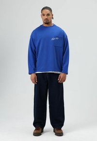 Blauwe sweatshirt met lange mouwen, een borstzak en witte stiksels, gecombineerd met losse, donkere spijkerbroeken en bruine wandelschoenen.
