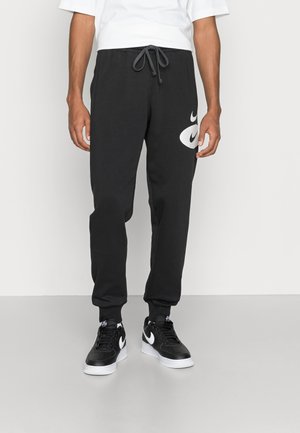 Man draagt zwarte Nike sweatpants met trekkoord in de taille en een wit swoosh-logo op de dij, gecombineerd met zwart-witte Nike sneakers.