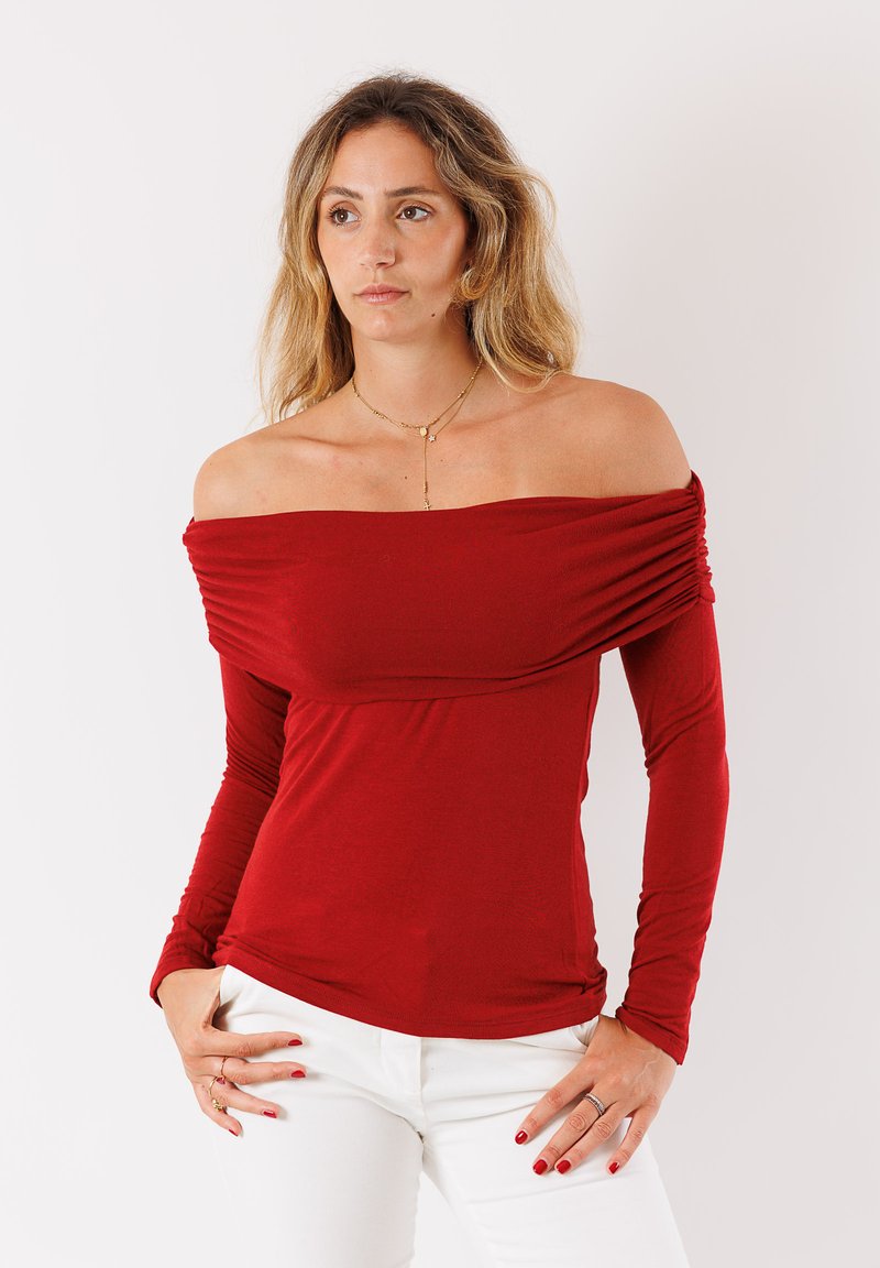 Top lungo rosso a spalla scoperta con dettaglio drappeggiato, realizzato in morbido tessuto. Indossato con pantaloni bianchi, che mettono in risalto il design aderente del top.