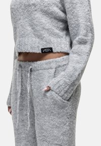 Close-up van een persoon in een zacht grijze gebreide lounge set met een cropped sweater en jogger broek, hand in de zak, met blote buik en gestructureerde stof.