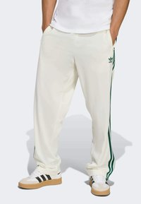 Pantalons de sport blancs avec des rayures latérales vertes. Fabriqués en tissu lisse. Équipés de poches et d'un logo brodé. Portés avec des chaussures blanches et noires.