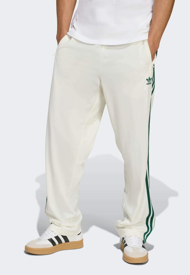 Pantalons de sport blancs avec des rayures latérales vertes. Fabriqués en tissu lisse. Équipés de poches et d'un logo brodé. Portés avec des chaussures blanches et noires.