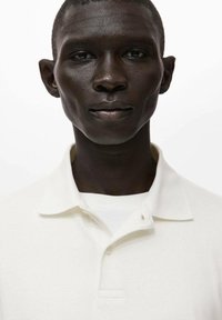 Weißes Poloshirt aus Baumwolle mit klassischem Kragen, Zwei-Knopf-Leiste und lockerer Passform. Die Textur wirkt weich und glatt.