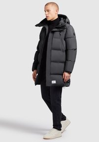 Must musta jope, millel on kapuuts, nelikantne disain, tõmbetaskud ja logo plekk. Kantud musta kõrge kaelusega pluusi ja tumedate teksade üle.