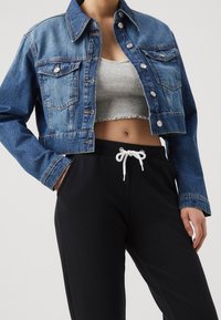 Personne portant une veste courte en jean bleu, un crop top gris côtelé et un pantalon noir avec un cordon blanc, une main dans la poche.