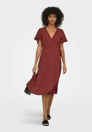 Kvinde med krøllet hår iført en rustrød omslagskjole til knæet, der holder en sort håndtaske og går i sorte blomstrede åbne tåhæl.